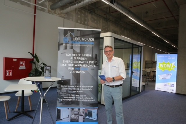 Ein Mann in weißem Hemd und grauer Hose steht neben einem Werbebanner in einem Gebäude, das wie ein Bürogebäude oder eine Veranstaltungshalle aussieht. Das Banner, überwiegend blau und weiß, wirbt für „Jörg Morach Energieberatung“ und enthält einen deutschen Text zur Auswahl der passenden Wärmepumpe. Der Mann hält eine kleine Broschüre oder Karte in den Händen und blickt den Betrachter leicht an. Im Hintergrund ist ein modernes Interieur mit Glaswänden, einem roten Feuerlöscher an der Wand und einem weiteren Banner mit farbenfrohen Grafiken in der Ferne zu sehen. Links neben dem Mann steht ein kleiner runder Tisch mit einer Pflanze.