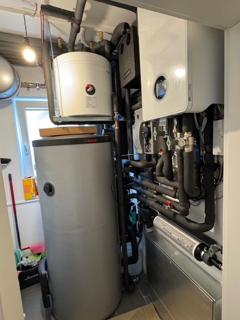 Ein beengter
Hauswirtschaftsraum, vollgestopft
mit diversen Komponenten der
Heizungs- und Wasseranlage. Links
steht ein hoher, zylindrischer,
grauer Bosch-Wassertank auf dem
Boden, darauf ein kleinerer,
weißer Zylinder. Hinter den Tanks
ist ein Fenster zu sehen, durch
das Tageslicht einfällt. Rechts an
der Wand hängen zwei weiße,
rechteckige Einheiten, vermutlich
ein Heizkessel und ein Bedienfeld,
unter denen zahlreiche schwarz
isolierte Rohre, Ventile und
Manometer verlaufen. Die Rohre
sind kunstvoll verlegt und
verbinden die verschiedenen
Komponenten. H
Glühbirne
SUG
hangt von der
und erhellt
den Raum.