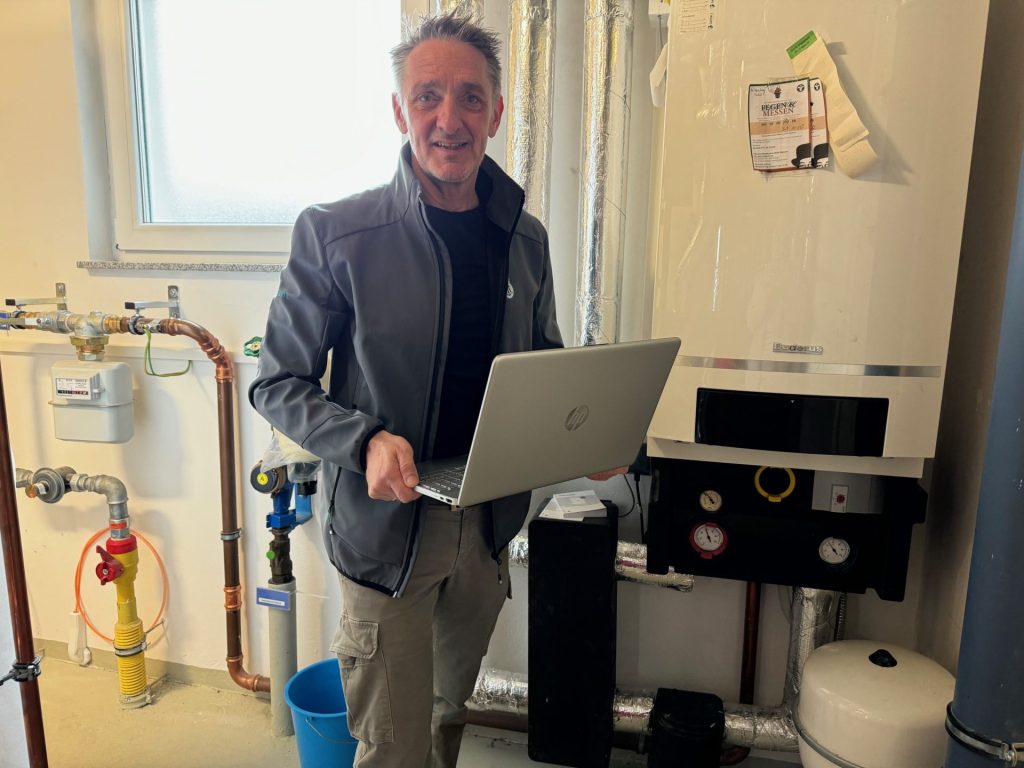 Ein lächelnder Mann mit ergrauendem Haar und Bart, bekleidet mit einer grauen Jacke und beigen Hosen, hält einen geöffneten silbernen Laptop in den Händen. Er steht in einem Hauswirtschaftsraum und blickt direkt in die Kamera. Links hinter ihm fällt helles Tageslicht durch ein Fenster, und ein Wasserzähler mit verschiedenen Rohren und Ventilen ist zu sehen. Rechts von ihm ist ein großer weißer Heizkessel an der Wand montiert, darunter befindet sich ein Bedienfeld mit Manometern. Isolierte Rohre verlaufen an der Wand über und hinter dem Mann. Unten links ist ein blauer Eimer teilweise zu erkennen.