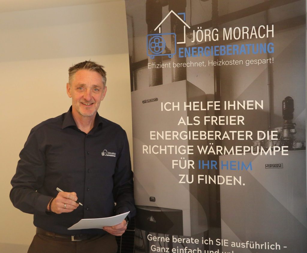 Ein Mann mit kurzem, ergrauendem Haar und Bart, bekleidet mit einem dunkelblauen Langarmhemd, steht lächelnd vor einem Werbebanner. Er hält ein Blatt Papier und einen Stift in der Hand und blickt direkt in die Kamera. Das Banner hinter ihm ist überwiegend weiß mit blauer und schwarzer Schrift. Es zeigt das Logo „Jörg Morach Energieberatung“ und den deutschen Text: „Ich helfe Ihnen als kostenloser Energieberater, die passende Wärmepumpe für Ihr Zuhause zu finden.“ Im Hintergrund des Banners ist ein Teil einer Wärmepumpe zu sehen.