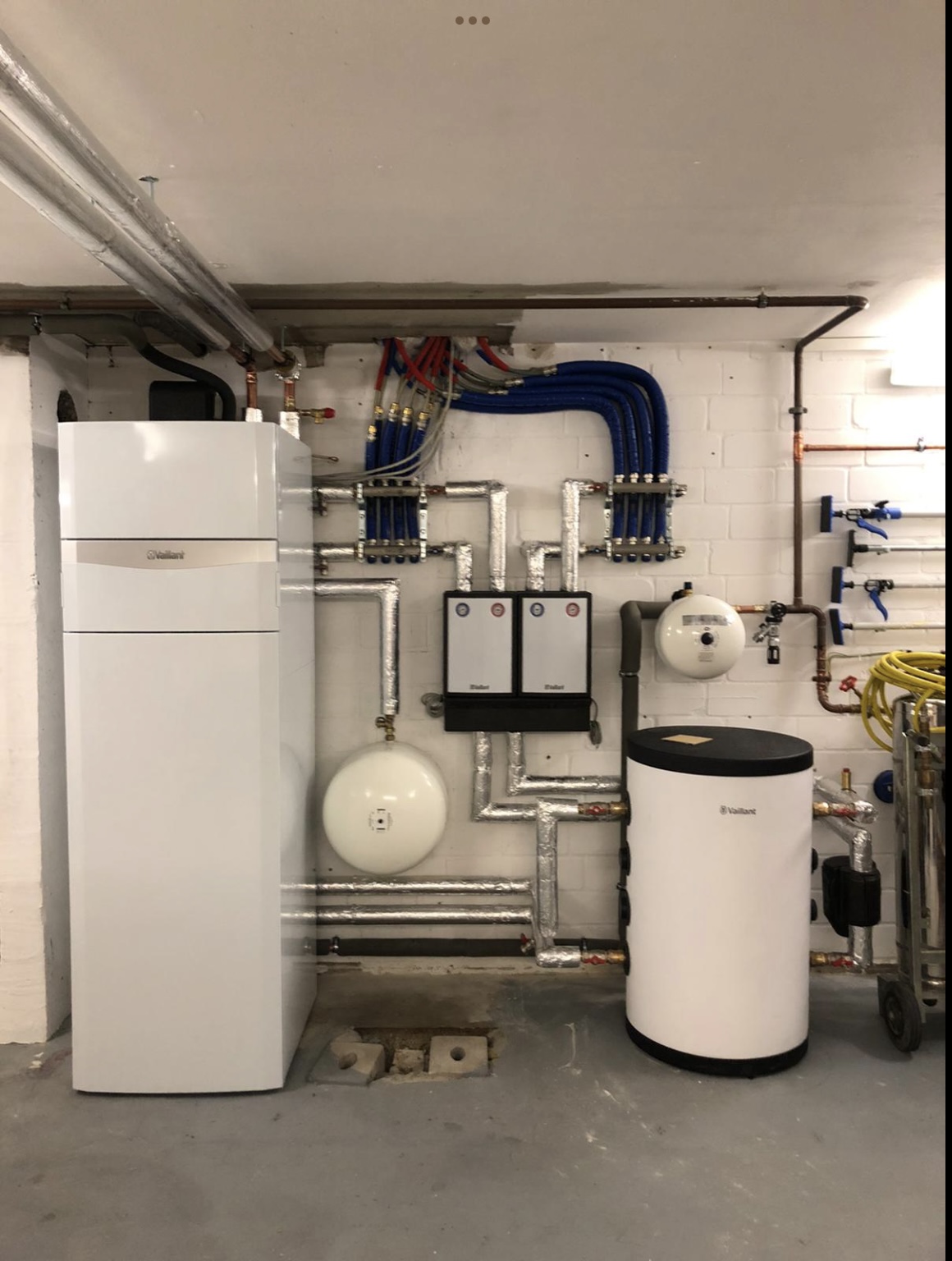 Ein sauberer, gut organisierter Hauswirtschaftsraum mit einer umfassenden Heizungs- und Wasseranlage. Links ragt eine hohe, elegante, weiße Wärmepumpe der Marke Vaillant empor. Rechts davon ist an einer weißen Ziegelwand ein komplexes Netzwerk aus silberisolierten Rohren, blauen und roten PEX-Rohren sowie verschiedenen Ventilen und Verteilern montiert. Zwei weiße, rechteckige Steuergeräte sind mittig an der Wand angebracht. Darunter sind zwei weiße, kugelförmige Ausdehnungsgefäße zu sehen, eines größer als das andere. Rechts im Bild steht ein niedrigerer, zylindrischer, weißer Vaillant-Wassertank mit schwarzem Deckel auf dem Boden. Weitere Kupfer- und weiße Rohre verlaufen entlang der rechten Wand. Der Boden ist aus Beton, und an der Decke sind Rohre und elektrische Leitungen sichtbar.
