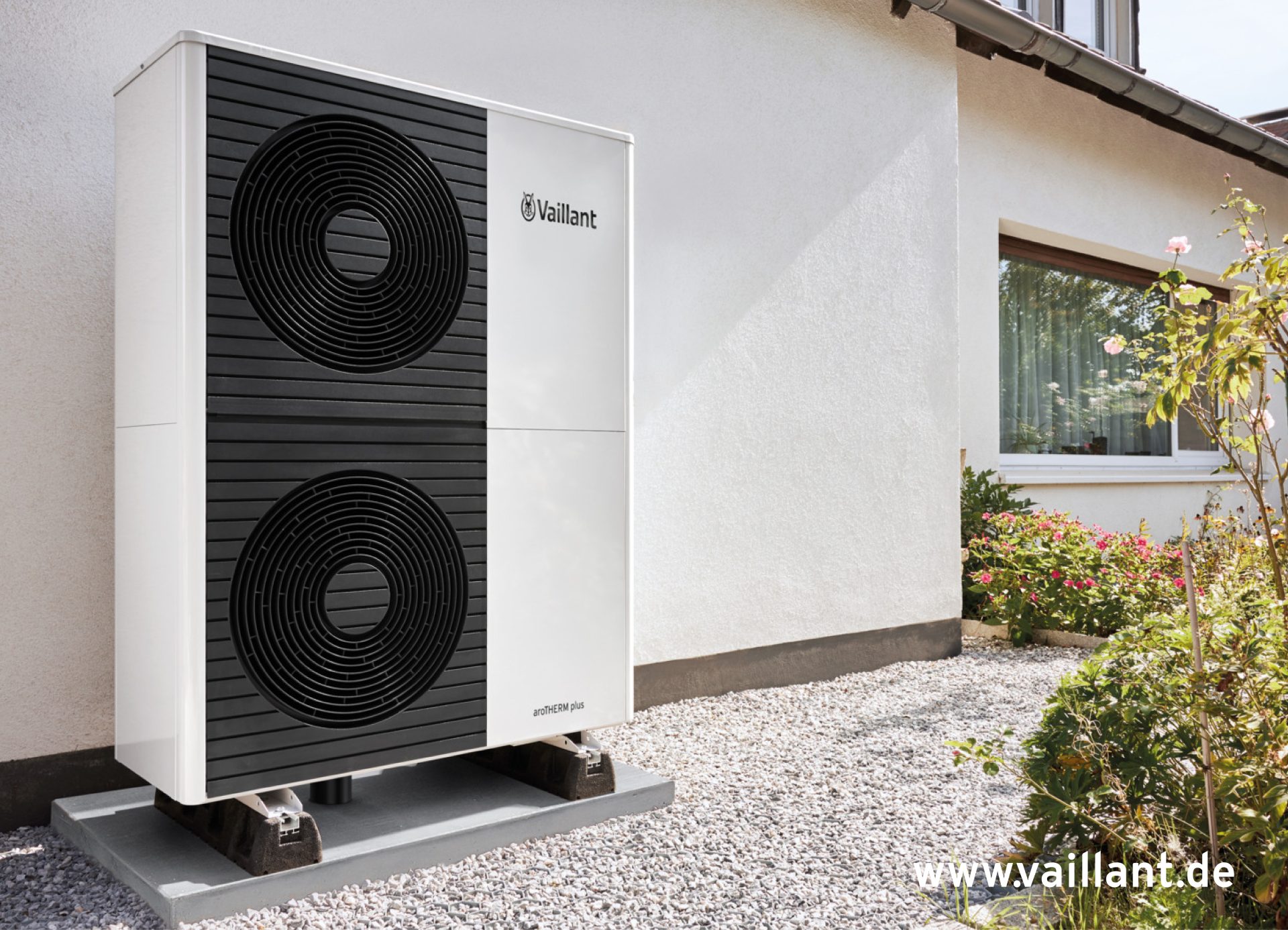 Eine elegante, weiße Vaillant-Wärmepumpe für den Außenbereich mit schwarzer Frontblende und zwei großen, runden Lüftergittern. Das Gerät steht auf dunklen Stützen auf einem grauen Sockel, eingebettet in eine Kiesfläche neben einem weiß getünchten Gebäude. Rechts davon befindet sich ein Garten mit grünen Sträuchern und einigen rosa Blüten. Am Gebäude ist ein Fenster mit weißen Rahmen zu sehen. Das Vaillant-Logo prangt prominent oben rechts am Gerät. Die Website „www.vaillant.de“ ist als Wasserzeichen unten rechts im Bild zu sehen.